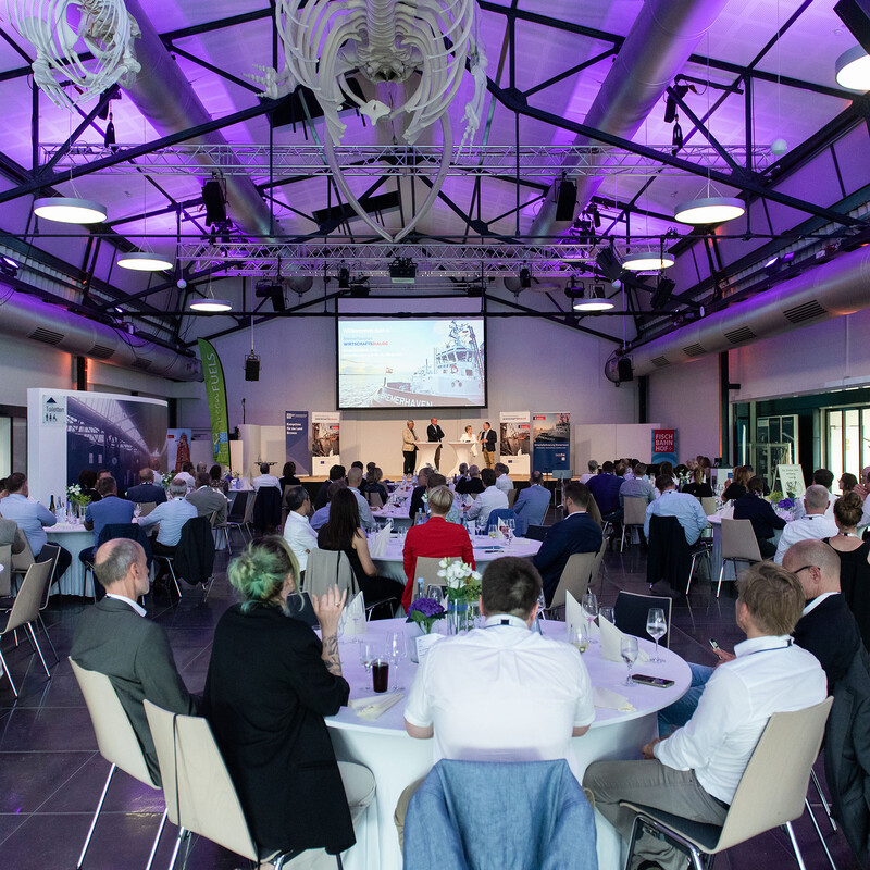 Ein großer Veranstaltungsraum mit Bühne auf der die Sprecher:innen stehen