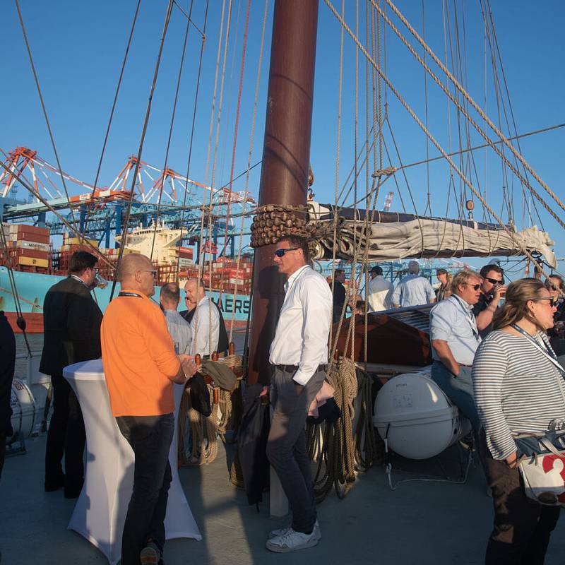 Viele Menschen an Deck eines Segelschiffes, im Hintergrund ein Containerschiff